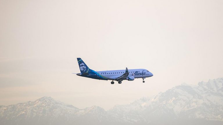 Blue and white Alaska Airlines plane landing with mountains in the background.