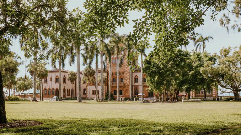 Tan colored Spanish-designed building sitting on the grounds with a large green yard and palm trees.