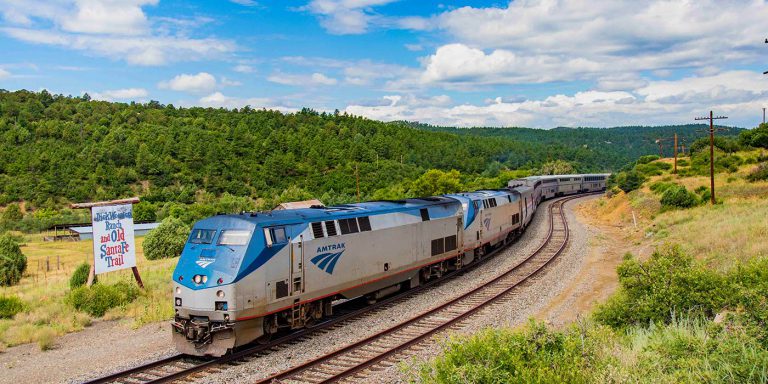 Silver and blue Amtrak train passing through green landscape of Raton Pass in New Mexico with the background of blue skies and white clouds.