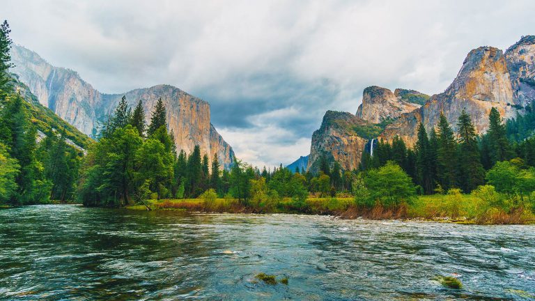 Towering rock peaks in Yosemite with green trees and river in the foreground.