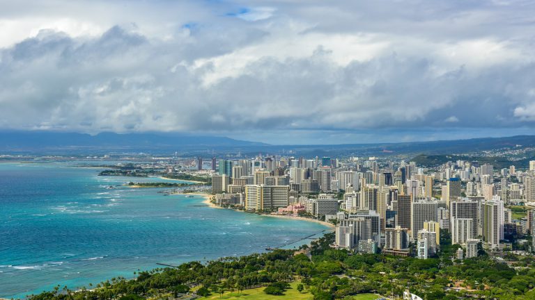 Blue water against a cloudy sky along with coast with tall buildings on the coast.