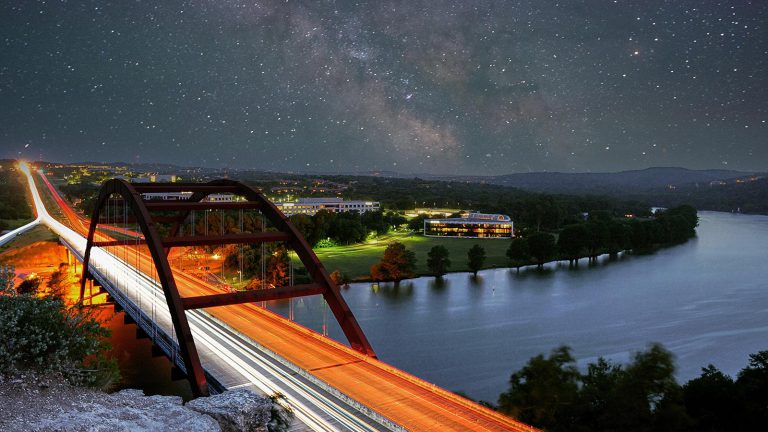 Blurred white and red car lights on a bridge against dark starry sky.