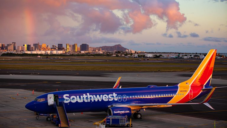 Blue, yellow, red plane sitting on runway with colorful clouds and Honolulu skyline behind it.