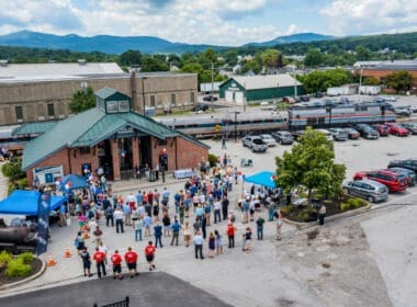 Amtrak train with train station in foreground and a large crowd standing in front of the train station.